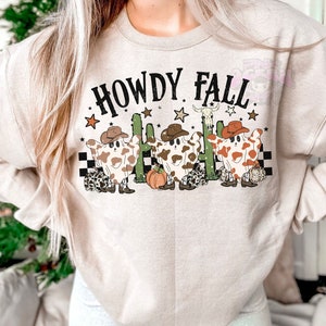 May include: Beige sweatshirt with the text "HOWDY FALL" in black. The graphic features three cow-shaped ghosts wearing cowboy hats, cacti, stars, and a pumpkin. The ghosts have brown and white spots and are wearing cowboy boots.
