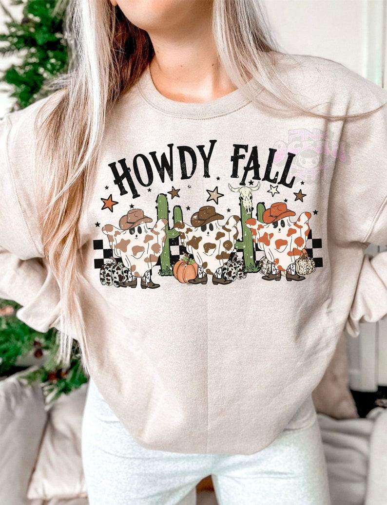 May include: Beige sweatshirt with the text "HOWDY FALL" in black. The graphic features three cow-shaped ghosts wearing cowboy hats, cacti, stars, and a pumpkin. The ghosts have brown and white spots and are wearing cowboy boots.
