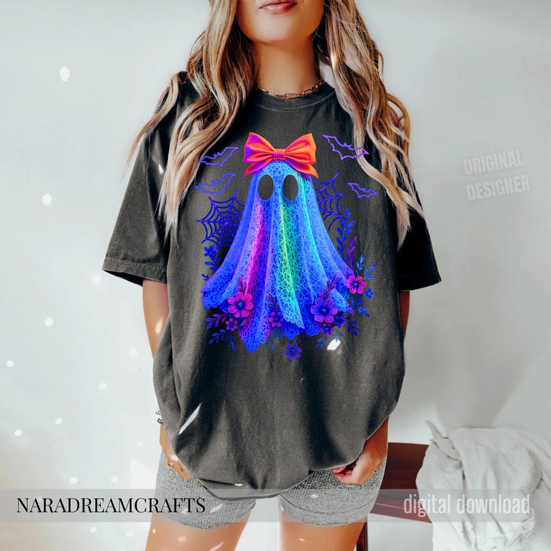 Peut inclure: T-shirt noir avec un motif de fant&ocirc;me color&eacute; orn&eacute; d'un n&oelig;ud, de fleurs et de chauves-souris. Le fant&ocirc;me est un m&eacute;lange vibrant de bleu, violet et vert. Le texte "NARADREAMCRAFTS" est en bas.