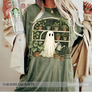 K&ouml;nnte beinhalten: Salbeigr&uuml;nes T-Shirt mit einer skurrilen Illustration eines Geistes, der Pflanzen in einem Gew&auml;chshaus gie&szlig;t. Das Design umfasst Topfpflanzen, eine Gie&szlig;kanne und den Text "NARADREAMCRAFTS" und "digital download".