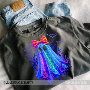 Peut inclure: T-shirt gris fonc&eacute; avec un motif de fant&ocirc;me color&eacute;, orn&eacute; d'un n&oelig;ud orange vif, de chauves-souris, de toiles d'araign&eacute;es et de fleurs violettes. Les textes "NARADREAMCRAFTS" et "digital download" sont visibles.