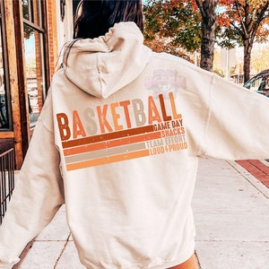 May include: A cream-colored hoodie with the word "Basketball" in brown, orange, and gray lettering. The text "Game Day Snacks Team Effort Loud & Proud" is printed in smaller lettering below the word "Basketball".