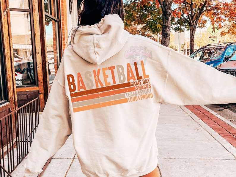May include: A cream-colored hoodie with the word "Basketball" in brown, orange, and gray lettering. The text "Game Day Snacks Team Effort Loud & Proud" is printed in smaller lettering below the word "Basketball".
