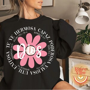 May include: Black sweatshirt with a pink flower graphic and the text "Dios" in the center. The flower has white petals and a white center. The text around the flower includes bible verses in Spanish.