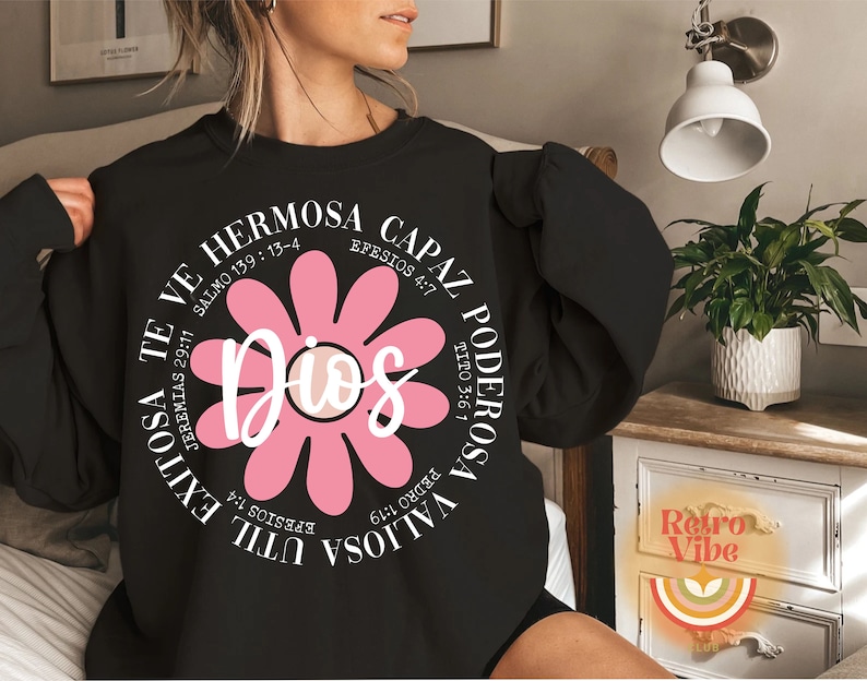 May include: Black sweatshirt with a pink flower graphic and the text "Dios" in the center. The flower has white petals and a white center. The text around the flower includes bible verses in Spanish.