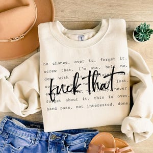 May include: Cream-colored sweatshirt with the phrase "fuck that" in a bold, black font. Surrounding text includes phrases like "no chance," "forget it," and "not interested." The sweatshirt is paired with blue denim shorts, a tan hat, and sandals.