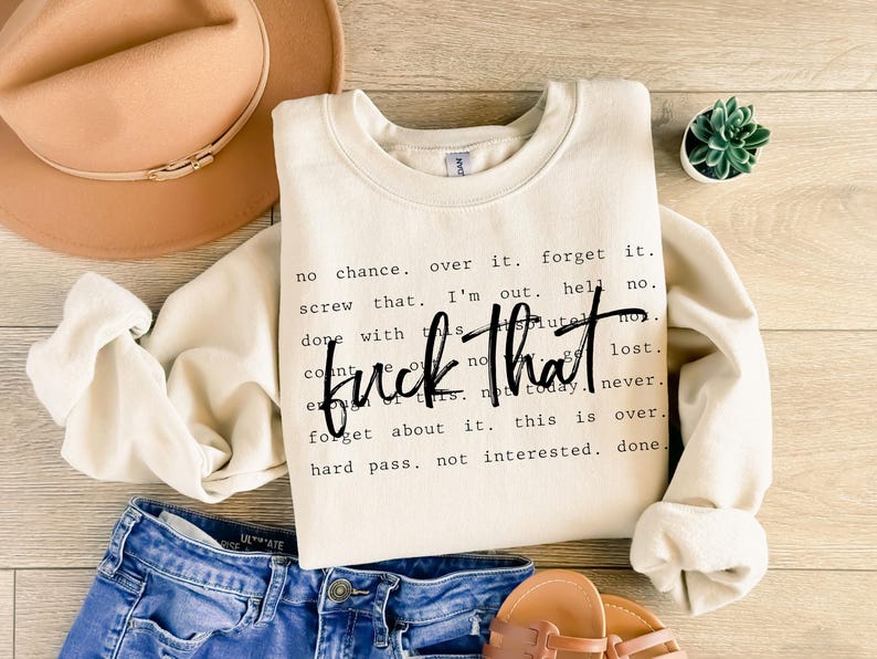 May include: Cream-colored sweatshirt with the phrase "fuck that" in a bold, black font. Surrounding text includes phrases like "no chance," "forget it," and "not interested." The sweatshirt is paired with blue denim shorts, a tan hat, and sandals.