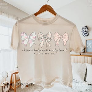 May include: Cream-colored t-shirt featuring three decorative bows in pastel colors. The bows have floral and patterned designs. The text "chosen holy and dearly loved" and "COLOSSIANS 3:12" are printed below the bows. The shirt is hanging on a wooden hanger.