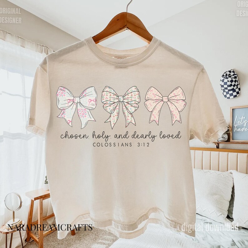 May include: Cream-colored t-shirt featuring three decorative bows in pastel colors. The bows have floral and patterned designs. The text "chosen holy and dearly loved" and "COLOSSIANS 3:12" are printed below the bows. The shirt is hanging on a wooden hanger.