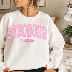 May include: White sweatshirt with the words "STRONGER" in pink block letters, with pink ribbon accents, above the words "THAN CANCER" in a smaller pink font. The sweatshirt is a crew neck style.