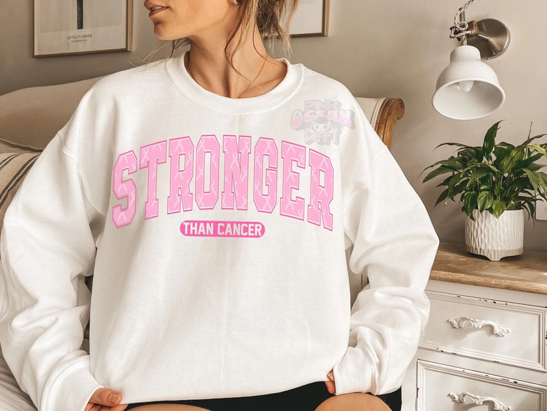 May include: White sweatshirt with the words "STRONGER" in pink block letters, with pink ribbon accents, above the words "THAN CANCER" in a smaller pink font. The sweatshirt is a crew neck style.