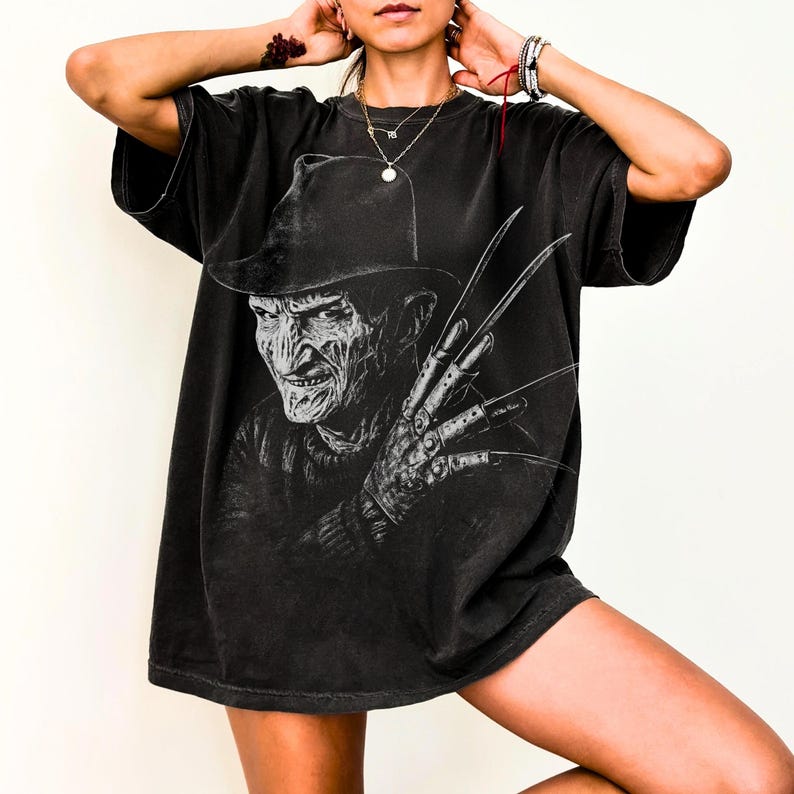 Puede incluir: Camiseta negra con un dise&ntilde;o gr&aacute;fico detallado de un personaje de pel&iacute;cula de terror con sombrero y guante con cuchillas. El dise&ntilde;o est&aacute; en tonos grises y negros, creando un aspecto vintage y desgastado. La camiseta tiene mangas cortas y un corte holgado.