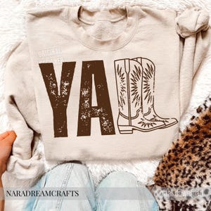 May include: Beige sweatshirt with the word "YA'LL" in distressed brown letters and a matching illustration of cowboy boots. The text "ORIGINAL DESIGNER" is printed above the word "YA'LL".