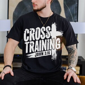 May include: Black t-shirt with the words "CROSS TRAINING" in a bold, white font, accompanied by a white cross graphic. The text "JOHN 3:16" is below the main text. The shirt is a casual wear item.