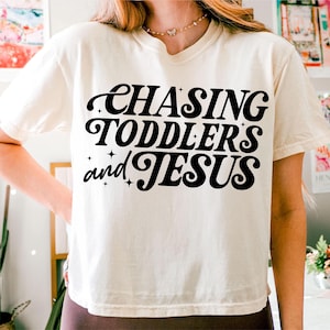 May include: A white cropped t-shirt with black text that reads "Chasing Toddlers and Jesus".