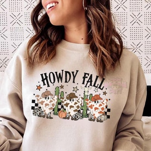 May include: Beige sweatshirt with the text "HOWDY FALL" in black. The graphic features three cartoon ghosts wearing cowboy hats, with a cactus, stars, and a pumpkin. The ghosts have brown and white cow-like patterns.