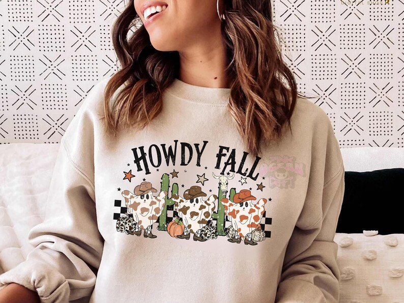 May include: Beige sweatshirt with the text "HOWDY FALL" in black. The graphic features three cartoon ghosts wearing cowboy hats, with a cactus, stars, and a pumpkin. The ghosts have brown and white cow-like patterns.