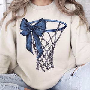 Peut inclure: Sweat-shirt cr&egrave;me avec une illustration d&eacute;taill&eacute;e d'un panier de basket et d'un grand n&oelig;ud bleu drap&eacute;. Le filet est finement dessin&eacute; et le n&oelig;ud ajoute un &eacute;l&eacute;ment d&eacute;coratif au th&egrave;me sportif. Le sweat-shirt est de couleur neutre.