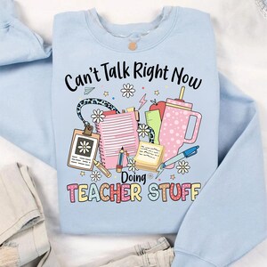 May include: Light blue sweatshirt with the text "Can't Talk Right Now Doing Teacher Stuff" in colorful letters. The text is surrounded by a variety of school supplies, including a pencil, a ruler, a notebook, a cup, and a flower.