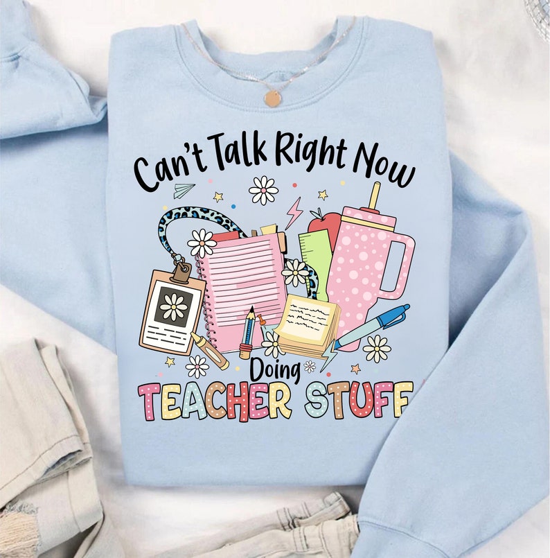 May include: Light blue sweatshirt with the text "Can't Talk Right Now Doing Teacher Stuff" in colorful letters. The text is surrounded by a variety of school supplies, including a pencil, a ruler, a notebook, a cup, and a flower.