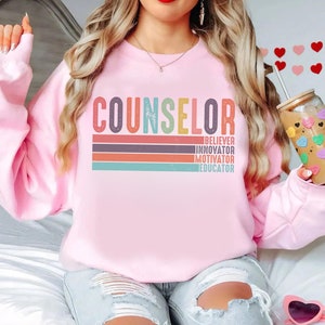 May include: Pink sweatshirt with the word "COUNSELOR" in rainbow colors and the words "BELIEVER, INNOVATOR, MOTIVATOR, EDUCATOR" in white below it.