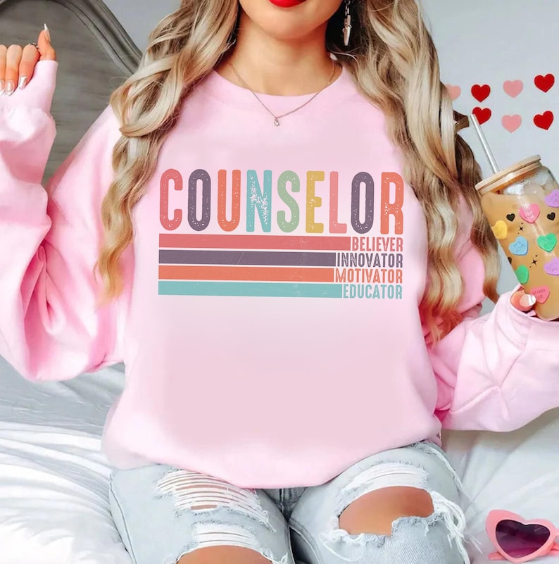 May include: Pink sweatshirt with the word "COUNSELOR" in rainbow colors and the words "BELIEVER, INNOVATOR, MOTIVATOR, EDUCATOR" in white below it.
