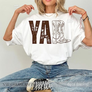 May include: White t-shirt with the brown text "YA" and a cowboy boot graphic. The shirt is a casual style, suitable for everyday wear. The design has a distressed, vintage look.