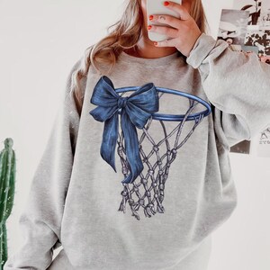 Peut inclure: Un sweat-shirt gris clair avec un motif de panier de basket-ball bleu et un n&oelig;ud bleu nou&eacute; sur le bord. Le filet est gris clair. Le sweat-shirt est de style d&eacute;contract&eacute;.