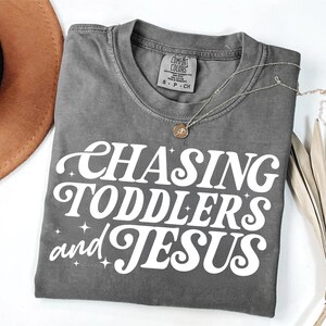 May include: A gray t-shirt with the text "Chasing Toddlers and Jesus" in white retro-style lettering.