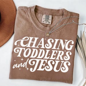 May include: A brown t-shirt with the text "CHASING TODDLERS and JESUS" in white with a retro font and sparkling stars.
