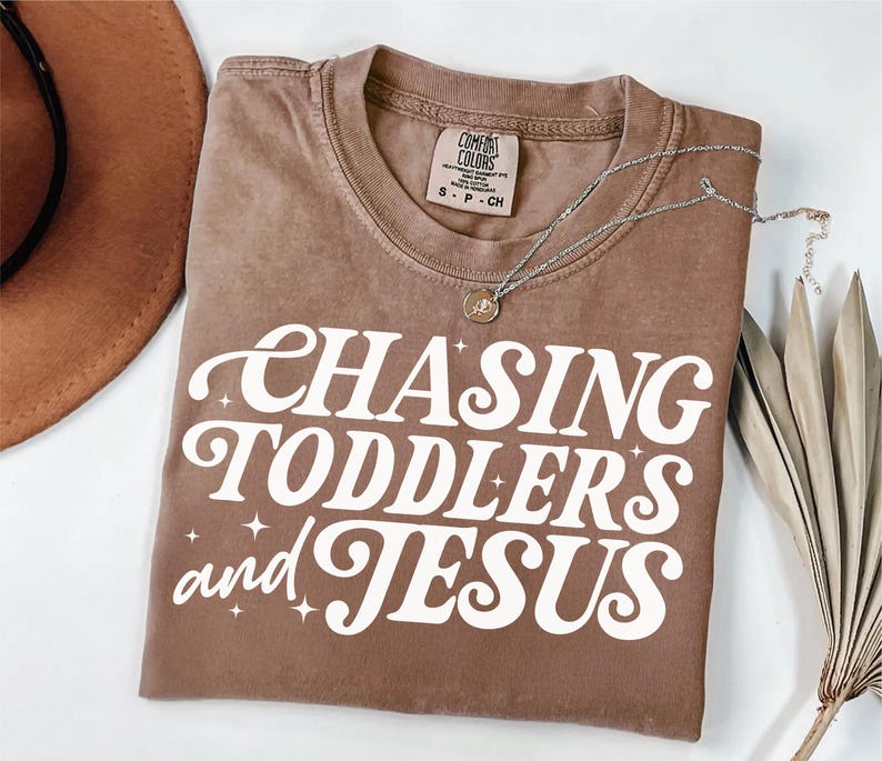 May include: A brown t-shirt with the text "CHASING TODDLERS and JESUS" in white with a retro font and sparkling stars.