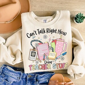 May include: A cream-colored sweatshirt with the text "Can't Talk Right Now Doing Teacher Stuff" in black lettering. The text is surrounded by colorful illustrations of school supplies, including a notebook, pencil, ruler, and a cup.