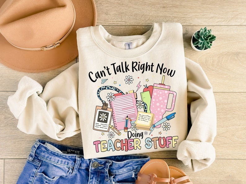 May include: A cream-colored sweatshirt with the text "Can't Talk Right Now Doing Teacher Stuff" in black lettering. The text is surrounded by colorful illustrations of school supplies, including a notebook, pencil, ruler, and a cup.