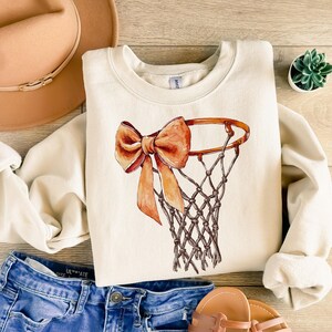 Op de afbeelding: Een cr&egrave;mekleurig sweatshirt met een bruine en oranje strik om een ​​basketbalring-afbeelding.