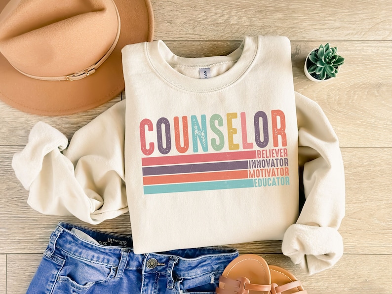 May include: A cream-colored sweatshirt with the word "COUNSELOR" in rainbow colors and the words "BELIEVER, INNOVATOR, MOTIVATOR, EDUCATOR" stacked vertically in a smaller font below.