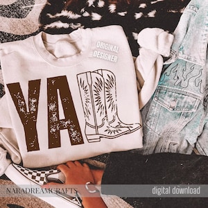 May include: A cream-colored sweatshirt with the word "YA" in distressed brown letters and a brown cowboy boot graphic. The words "ORIGINAL DESIGNER" are printed on the top right. A denim jacket and checkered shoes are also visible.