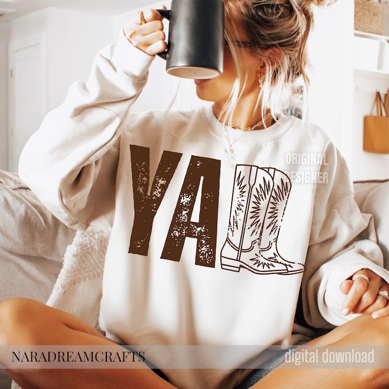 May include: Cream-colored sweatshirt with the word "YA" in brown block letters and a graphic of a cowboy boot. The words "ORIGINAL DESIGNER" are printed on the right side of the sweatshirt.