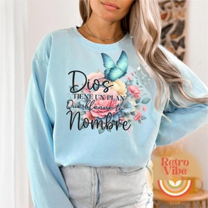 May include: Light blue sweatshirt with the text "Dios tiene un plan que lleve tu nombre" in black script. A floral design with pink, yellow, and blue flowers and a turquoise butterfly is on the right side. The Retro Vibe Club logo is in the bottom right.