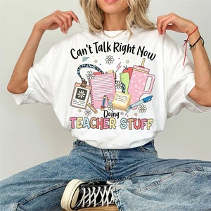 May include: White t-shirt with the text "Can't Talk Right Now Doing Teacher Stuff" and a graphic of school supplies, including a pencil, notebook, and a cup.