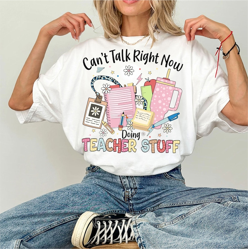 May include: White t-shirt with the text "Can't Talk Right Now Doing Teacher Stuff" and a graphic of school supplies, including a pencil, notebook, and a cup.