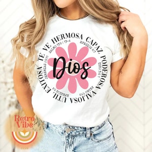 May include: White t-shirt with a pink flower graphic and the word "Dios" in the center. The flower has a circle around it with text in Spanish that translates to "You are valuable, strong, capable, beautiful, and God loves you".