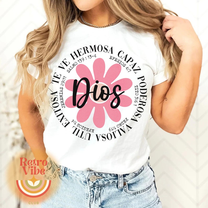 May include: White t-shirt with a pink flower graphic and the word "Dios" in the center. The flower has a circle around it with text in Spanish that translates to "You are valuable, strong, capable, beautiful, and God loves you".