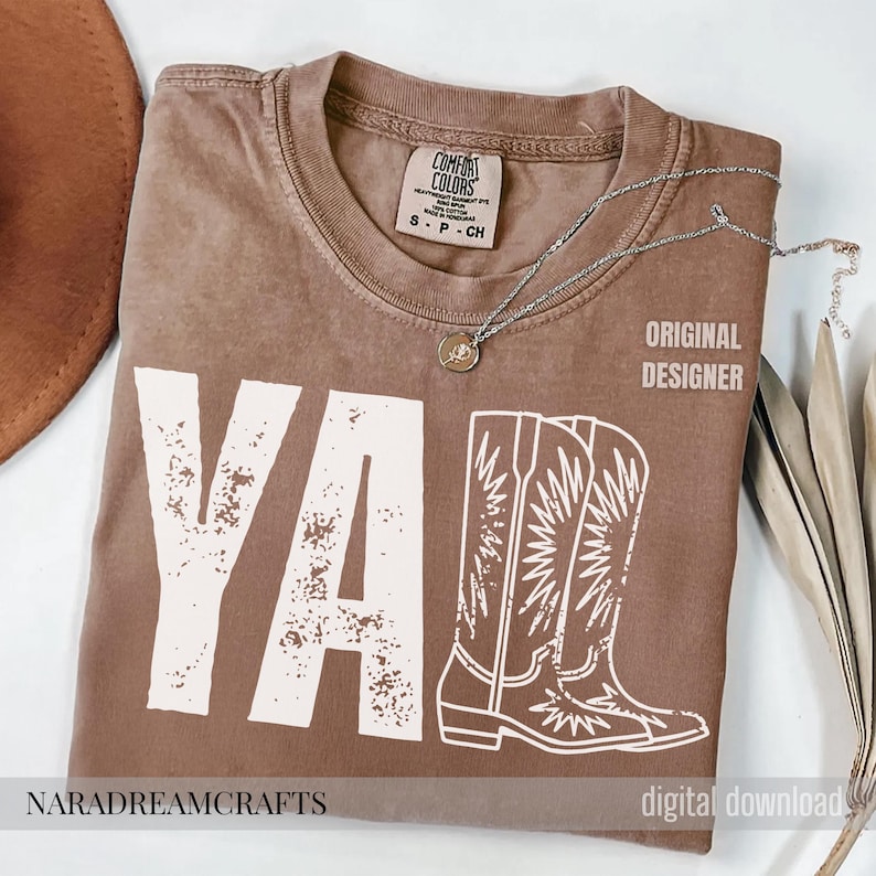 May include: A brown Comfort Colors t-shirt with the word "YA" and a pair of cowboy boots printed in white. The text "ORIGINAL DESIGNER" is printed on the right side. A silver necklace with a pendant is on the shirt.