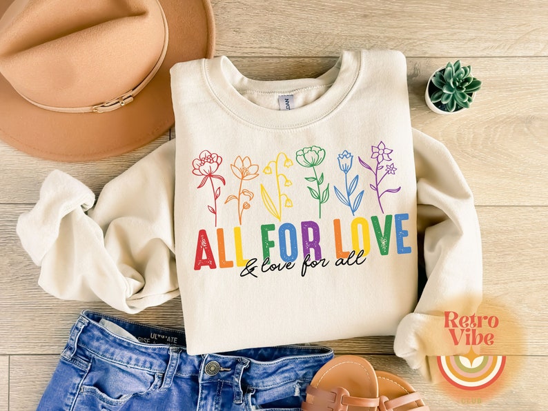 Puede incluir: Una sudadera de color crema con un dise&ntilde;o de arco&iacute;ris de flores y el texto "ALL FOR LOVE & love for all".