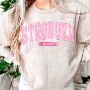 May include: Beige sweatshirt with the words "STRONGER THAN CANCER" in pink block letters. The word "STRONGER" is filled with pink ribbons. The text is centered on the front of the sweatshirt.