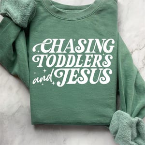 May include: A green sweatshirt with the text "CHASING TODDLERS and JESUS" in white retro-style lettering.