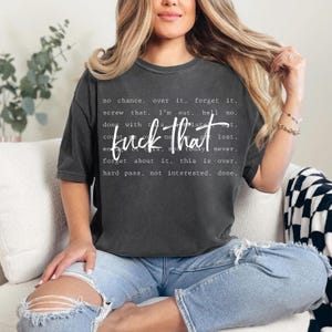 May include: Dark gray t-shirt with the phrase "fuck that" in a bold, white font. Surrounding the phrase are additional white words and phrases. The shirt is worn with blue jeans.