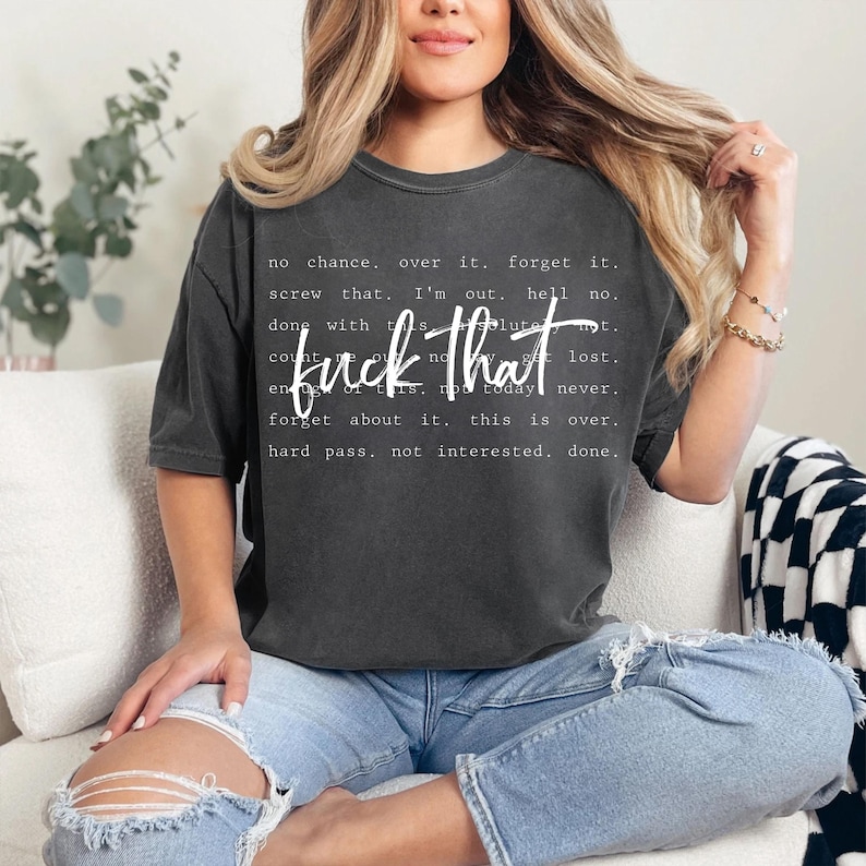 May include: Dark gray t-shirt with the phrase "fuck that" in a bold, white font. Surrounding the phrase are additional white words and phrases. The shirt is worn with blue jeans.