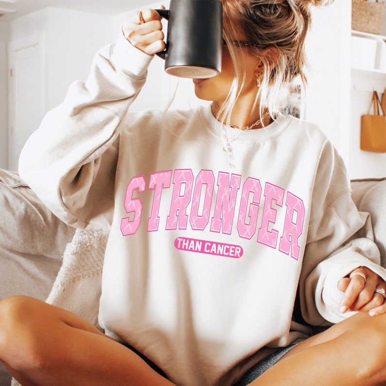 May include: A cream-colored sweatshirt with the text "STRONGER than CANCER" in pink block letters. The word "STRONGER" has a pink ribbon pattern. The sweatshirt is a casual style, suitable for everyday wear.