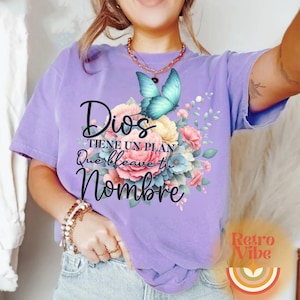 May include: A lilac-colored t-shirt featuring a floral design with a blue butterfly. The text "Dios tiene un plan que lleva tu nombre" is printed on the shirt. The design includes pink, yellow, and blue flowers.